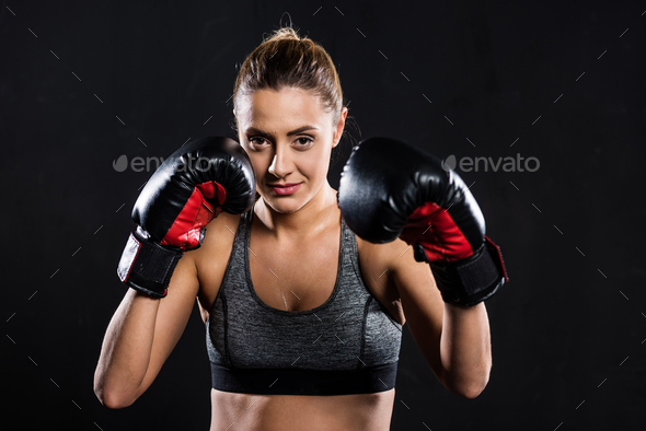 attractive young sportswoman in boxing gloves looking at camera isolated on black