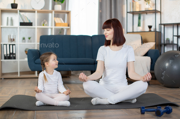 Happy mother with little daughter meditating, sitting in easy seat pose on mat at home