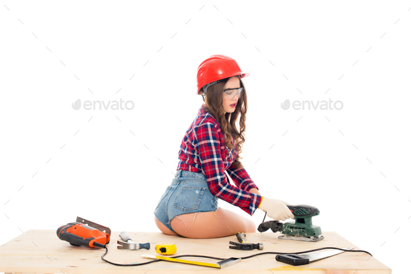 sexy girl in hardhat working with grind tool on wooden table with tools, isolated on white