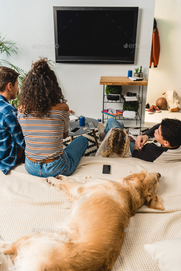 four teens watching tv and dog lying on bed