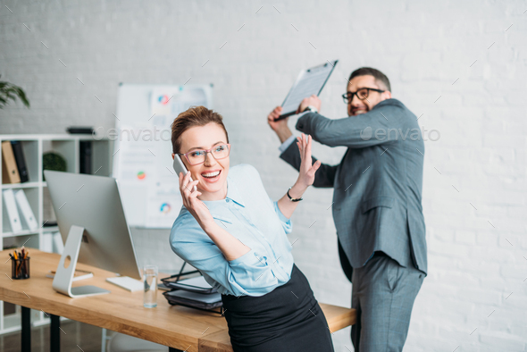 businessman get ready to hit his colleague with clipboard while she talking by phone and annoying