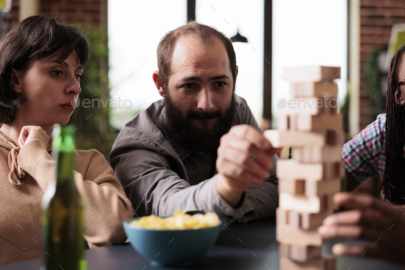 Focused man pulling wood block from tower structure while playing society games with friends.