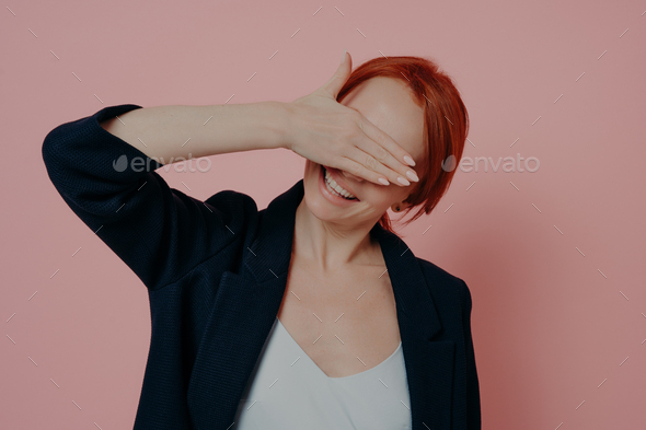 Cute positive ginger woman holding hand in front of face, covering eyes in anticipation for surprise