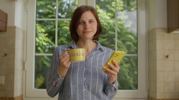 30s Woman Drinking Coffee of Yellow Cup with 'Dream' Sing Using Mobile Phone Standing in the Kitchen alt