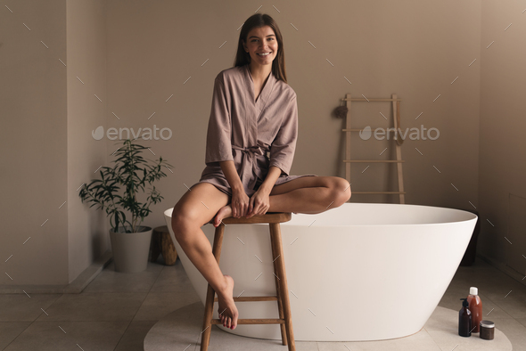 Beautiful woman sitting on high stool in bathroom going to take bath,  filling bathtub with water Stock Photo by damirkhabirov