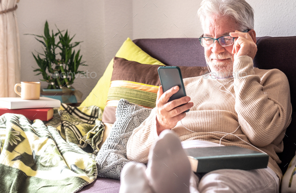 Handsome white-haired elderly man with earphones lying down on sofa at home using phone
