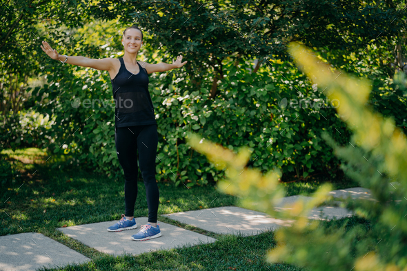 woman keeps arms sideways being in good mood exercises outdoors wears sportsclothes