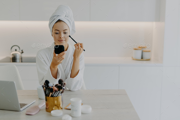 Young refreshed lady applies face powder with cosmetic brush looks at herself in mirror