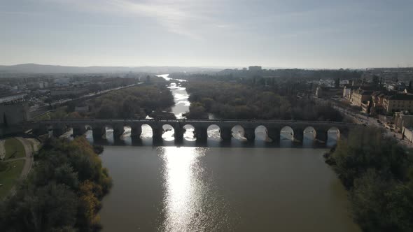 Aerial drone view of medieval Roman bridge in background with sunlight reflecting over Guadalquivir