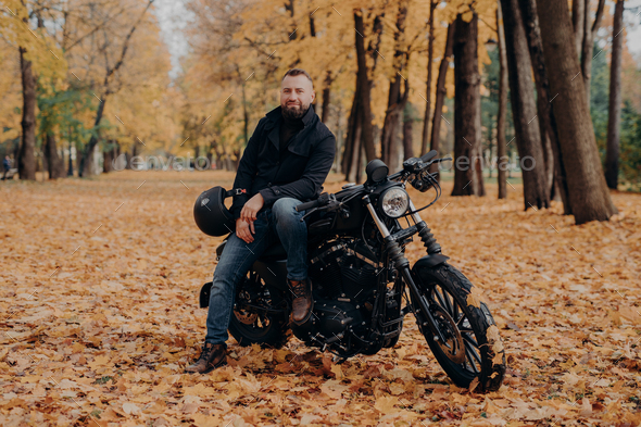 Bearded biker poses on own motorcycle, holds helmet, rides motorbike, poses outdoor in park