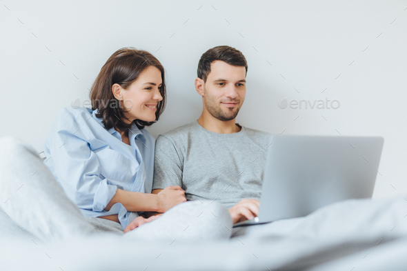 Affectionate female and male have joy together, watch film in bedroom on laptop