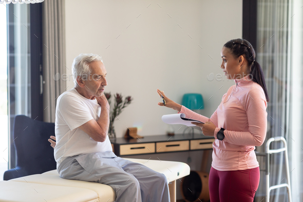 Biracial female physiotherapist with clipboard discussing with caucasian senior man at home - Stock Photo - Images