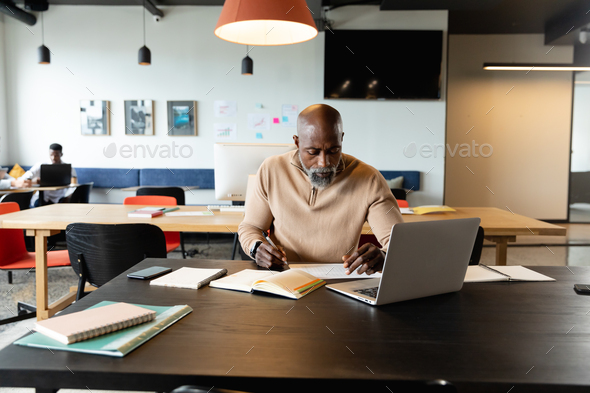 African american mature businessman writing in diary while sitting with at laptop in creative office - Stock Photo - Images