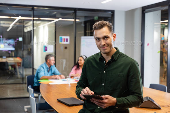 Portrait of confident young caucasian businessman with tablet pc while colleagues in background - Stock Photo - Images