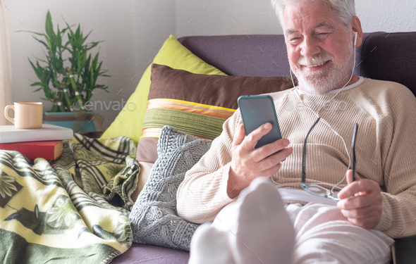 Handsome white-haired elderly man with earphones lying down on sofa at home using smart phone