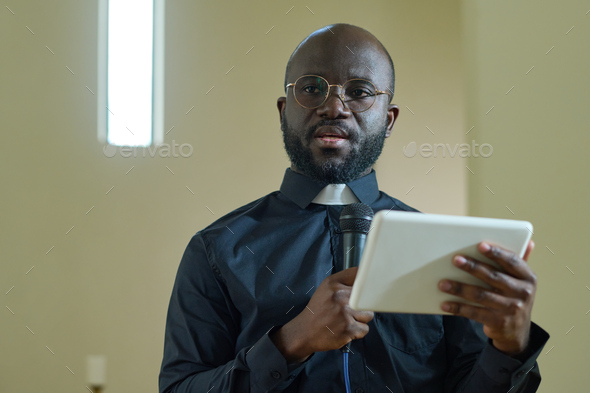 Young confident black man with microphone and digital tablet making speech