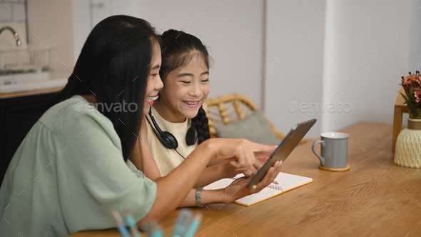 Smiling preteen girl sitting with mother in kitchen and having learning online at virtual class.