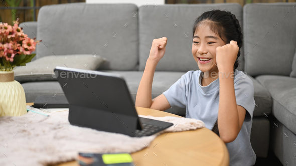 Happy asian child sitting in living room and learning online at virtual class via computer tablet.