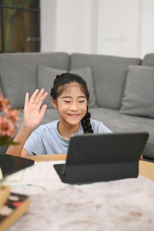 Cute girl having learning online at virtual class on computer tablet while sitting in living room.