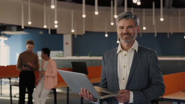 Portrait of Handsome Businessman Looking at the Camera Holding Laptop Standing in the Office alt