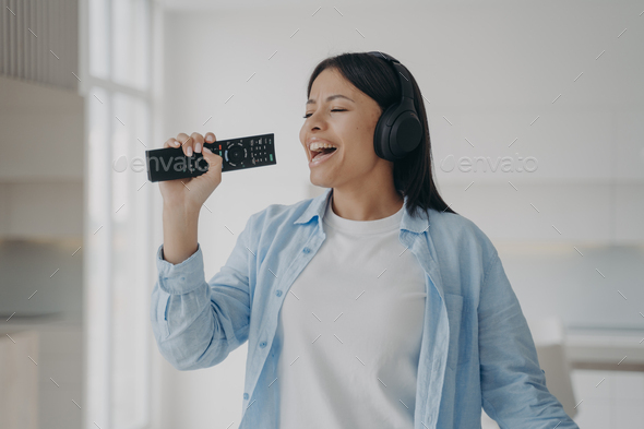Excited woman in headphones sing to imaginary microphone, TV remote control, listen to music at home