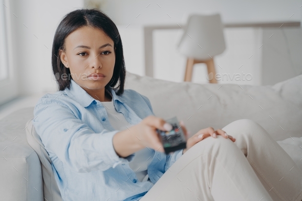 Bored female holding tv remote control change channels, watching television sitting on couch at home