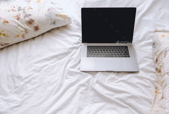 Laptop on a white bed with floral pillows. Work at home, concept of remote work freelancer