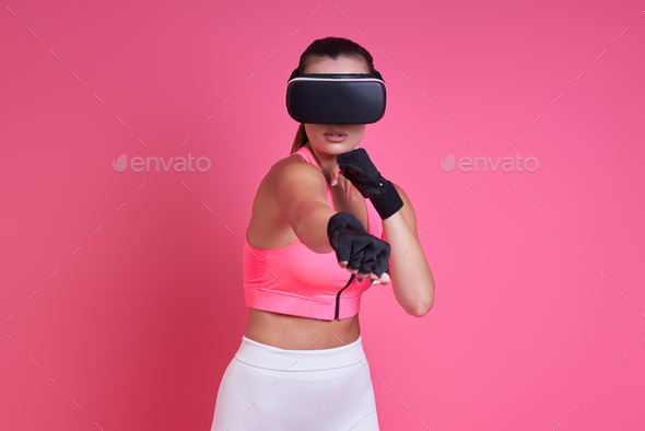Young woman in virtual reality glasses boxing against pink background - Stock Photo - Images