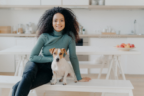 housewife with Afro haircut, sits at bench with pedigree dog, have fun and look directly at camera