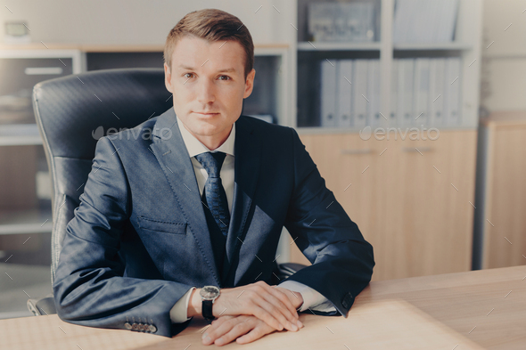 Professional male banker sits in cabinet, wears formal clothes, has confident expression