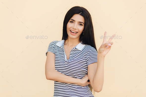 Portrait of cheerful brunette female with joyful expression, makes peace sign with two fingers