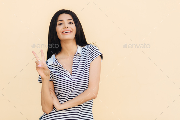 Portrait of cheerful brunette female shows peace gesture, keeps two fingers raised