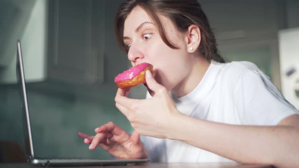 A Young Woman Eats a Pink Doughnut and Looks at a Laptop in Her Kitchen. Remote Work and Home
