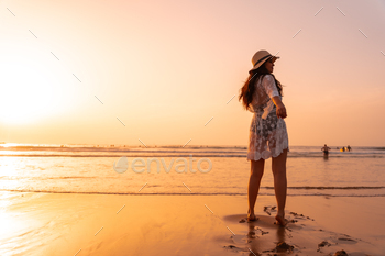 A woman in the sunset in a white dress at the sea in summer with a hat