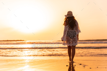 A woman at sunset in a white dress with a hat walking by the sea at low tide