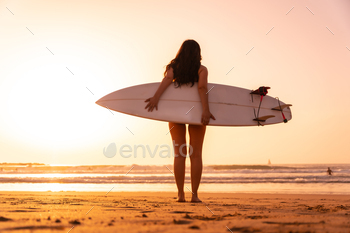 Portrait, a surfer woman on a beach at sunset with the surfboard looking at the sea