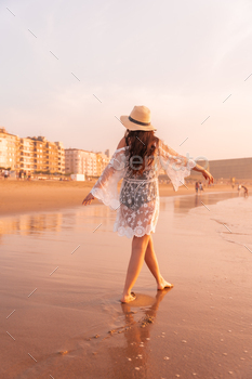Portrait of a woman at sunset in a white dress with a hat walking by the sea at low tide