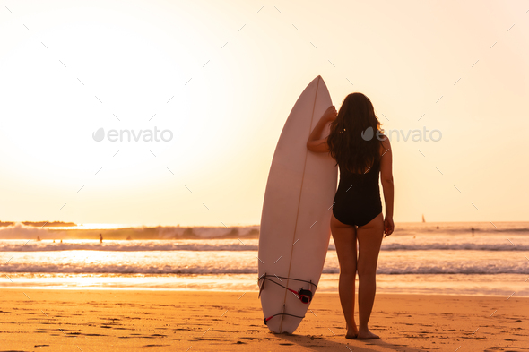 Surfer woman looking at the waves on a beach at sunset next to the surfboard
