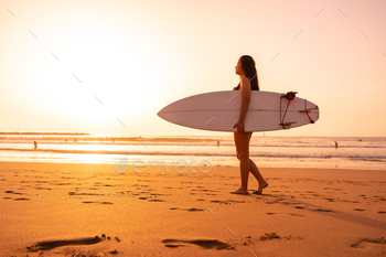 A surfer woman on a beach at sunset with the surfboard walking towards the sea