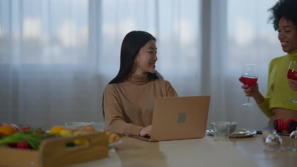 Overworking Asian Young Slim Woman Sitting with Laptop in Kitchen As African American Friend alt