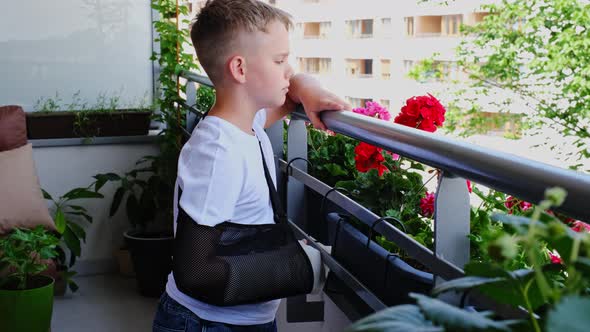 Sad Child with a Broken Arm in a Cast Stands on the Balcony and Waves to Someone alt