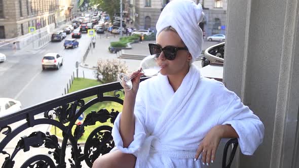 Young Woman in a Bathrobe Drinks Champagne on the Balcony