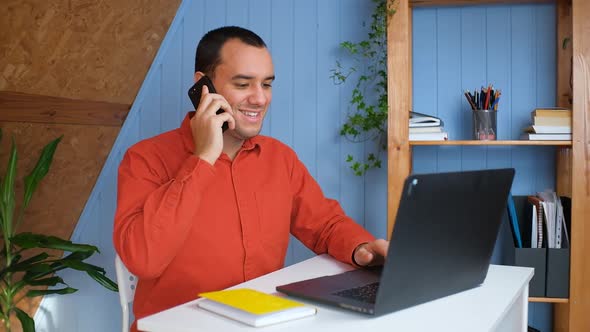 Smiling Young Business Man Professional Talking on Phone Using Laptop Sit at Home Office Desk alt