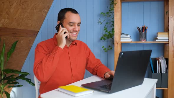 Smiling Young Business Man Professional Talking on Phone Using Laptop Sit at Home Office Desk