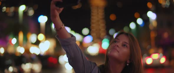A young woman taking a selfie in the middle of the street, bokeh lights alt