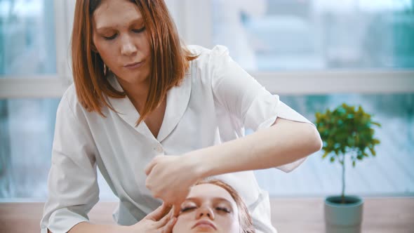 Massage - Masseuse in White Coat Kneading the Chin Area To a Young Woman Using Her Palms alt
