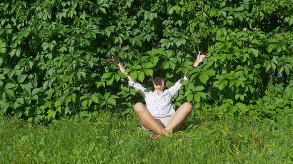 Wide Smiling Woman Sits on Grass Lawn Near Wall All Over Covered with Thick Ivy alt