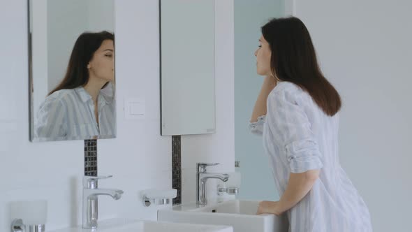 Attractive  young brunette woman in long shirt looking at herself in the mirror and fixing hair alt