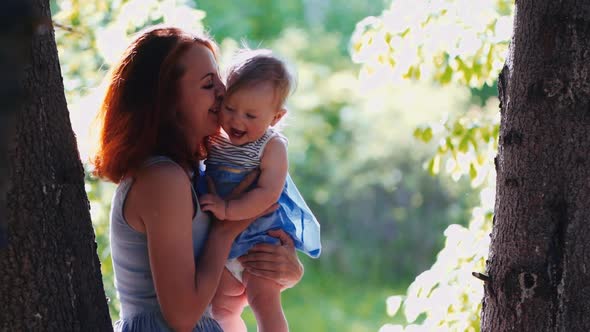 baby and mother are playing standing in the forest by a tree trunk in backlight alt
