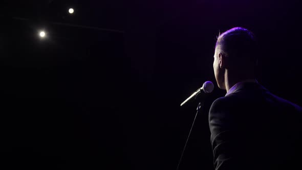 Man Stand Up Comedian Speaking Jokes in Micropphone Stands on Stage, Back View. alt
